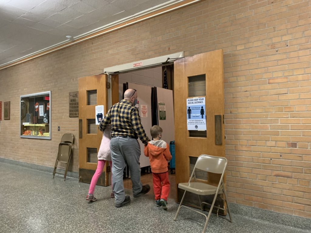Dad in plaid shirt walking with young daughter and son through a door in to the polling place.